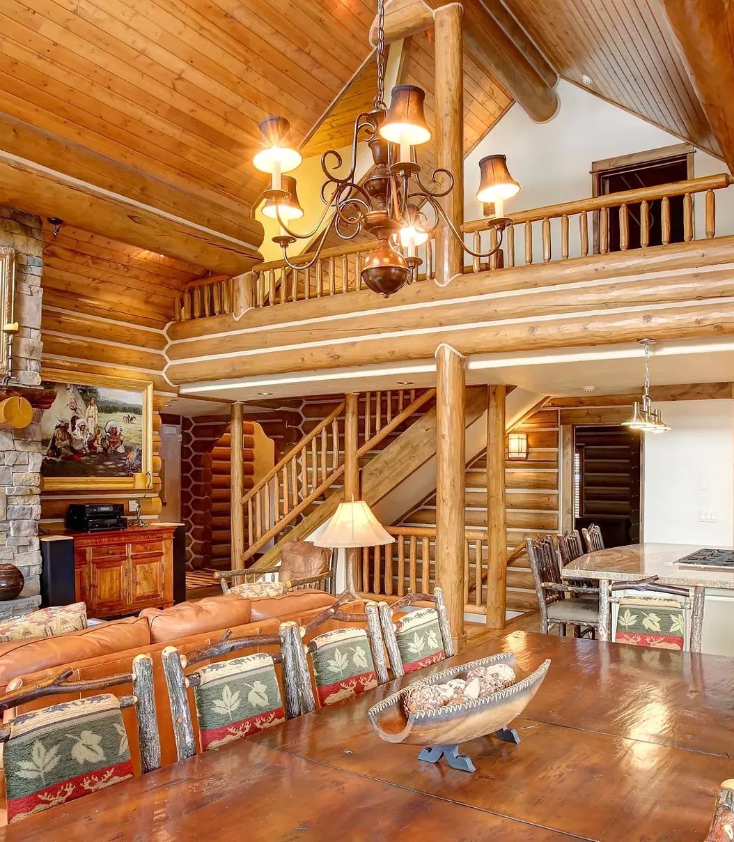 Rustic log cabin living room with wooden loft, chandelier, leather sofa, and stone fireplace