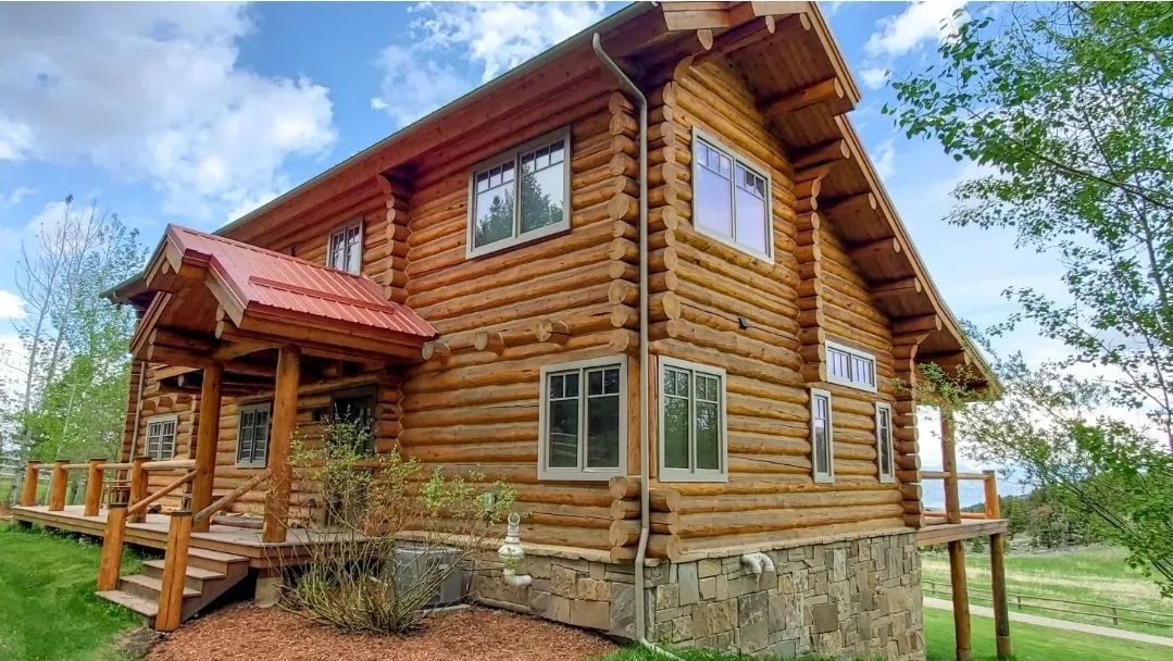Rustic wooden cabin with a red roof and stone foundation in a grassy yard under a blue sky