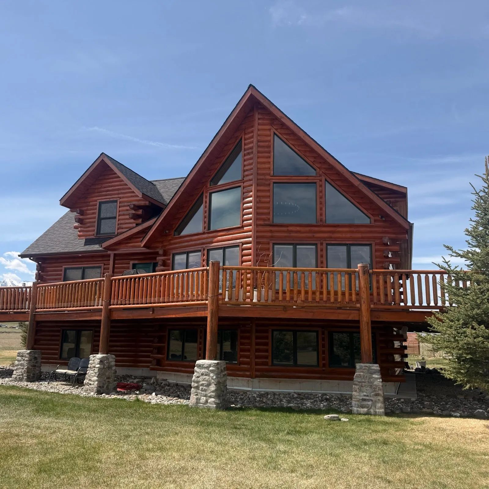 Rustic red log cabin with a wooden deck, stone pillars, and large windows under a blue sky.