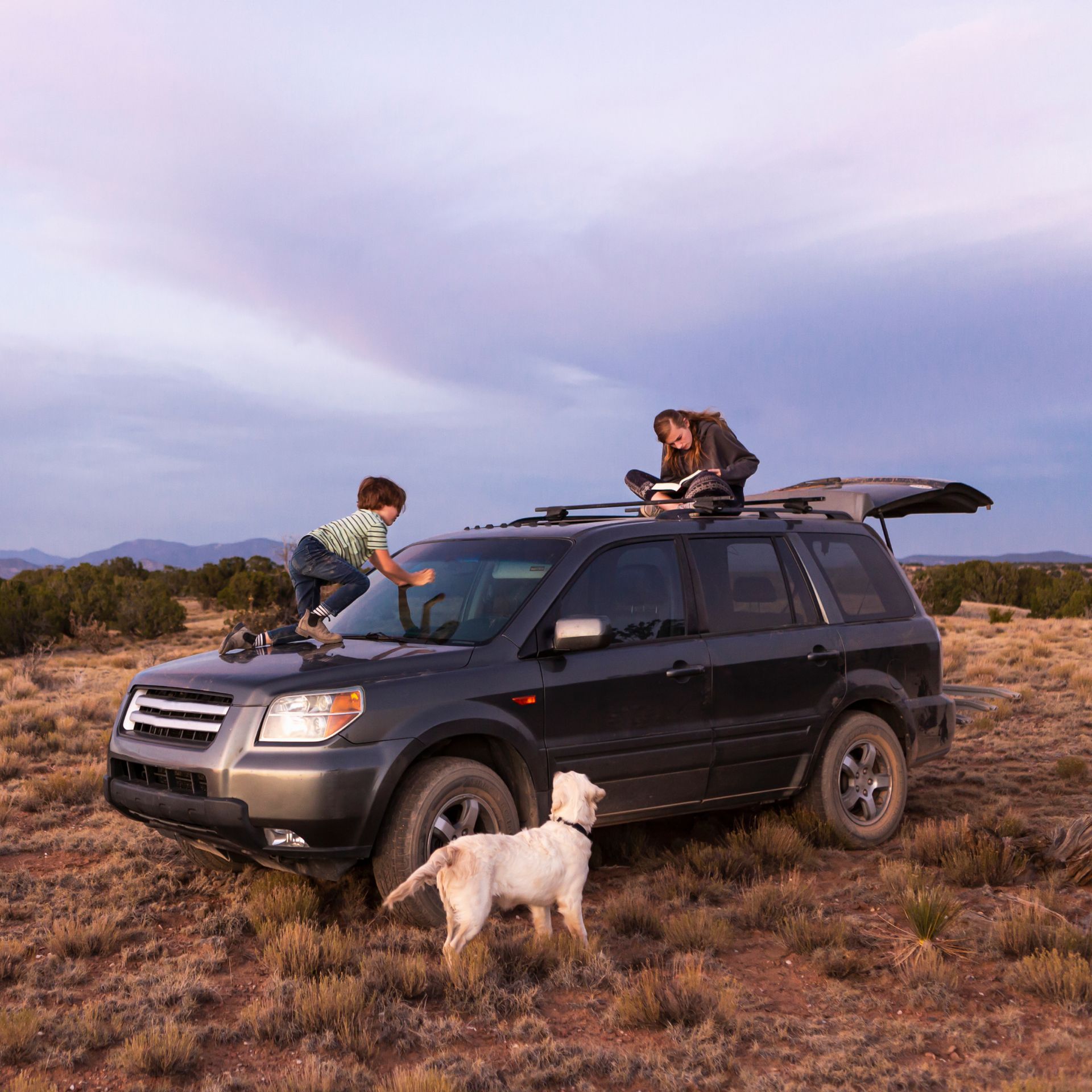 A boy and a woman are sitting on top of a suv with a dog standing next to it