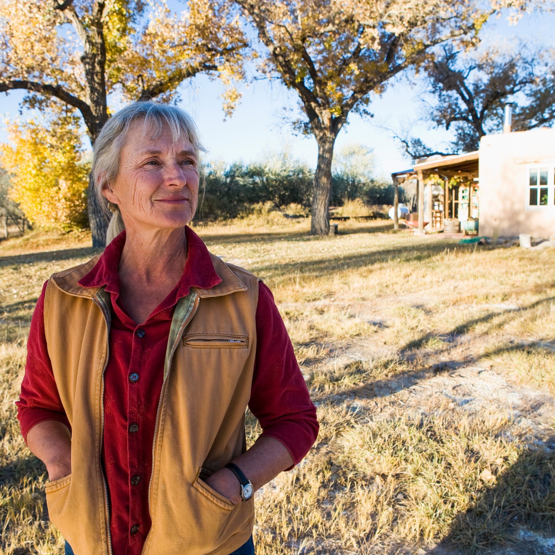 A woman wearing a vest and a red shirt is standing in front of a house.