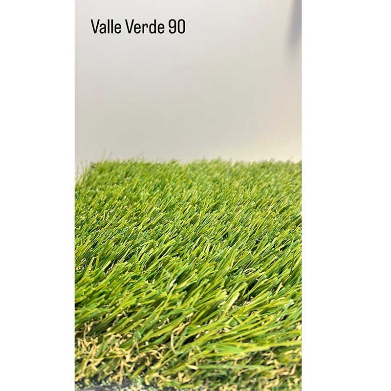 Primer plano de césped artificial de color verde intenso contra una pared blanca lisa con el texto «Valle Verde 90».
