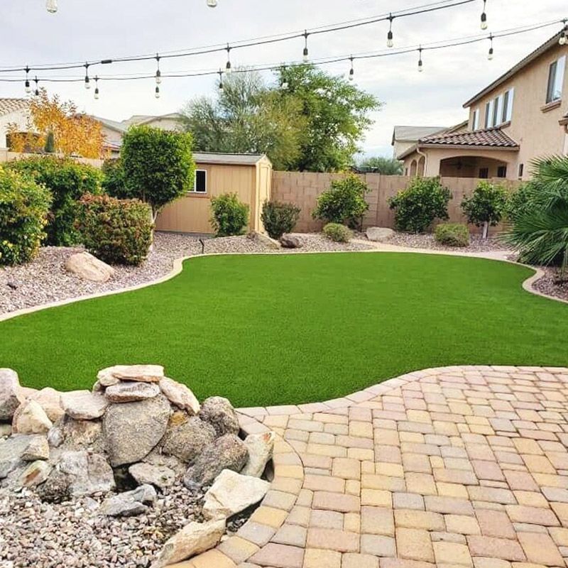 Backyard with green turf, stone patio, string lights, and landscaped garden beside a house