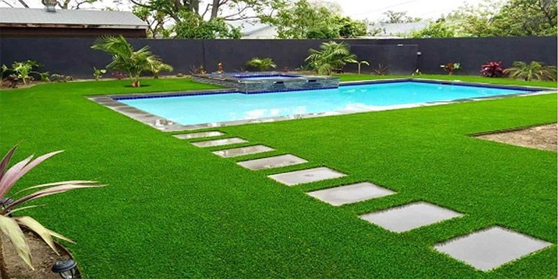 Patio trasero con césped verde, piedras de paso y una piscina turquesa rodeada de una valla oscura