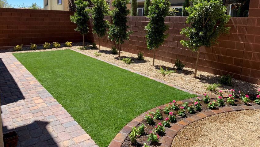 Patio ajardinado con césped artificial, sendero de ladrillo, parterre y árboles pequeños junto a un muro de ladrillo rojo