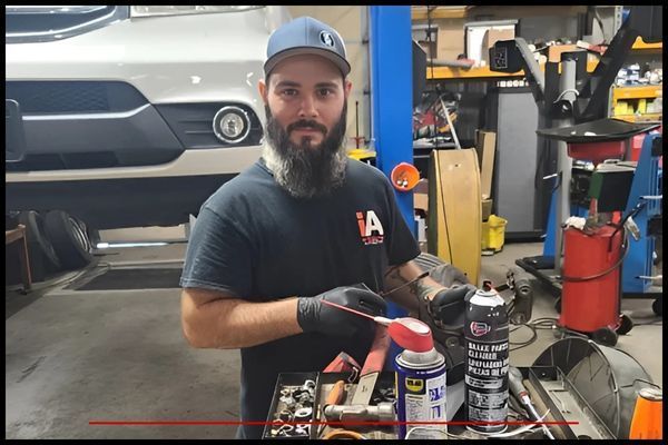 Mechanic in a garage sprays a part with a can of lubricant. He wears a hat, gloves, and a t-shirt. | Integrated Automotive