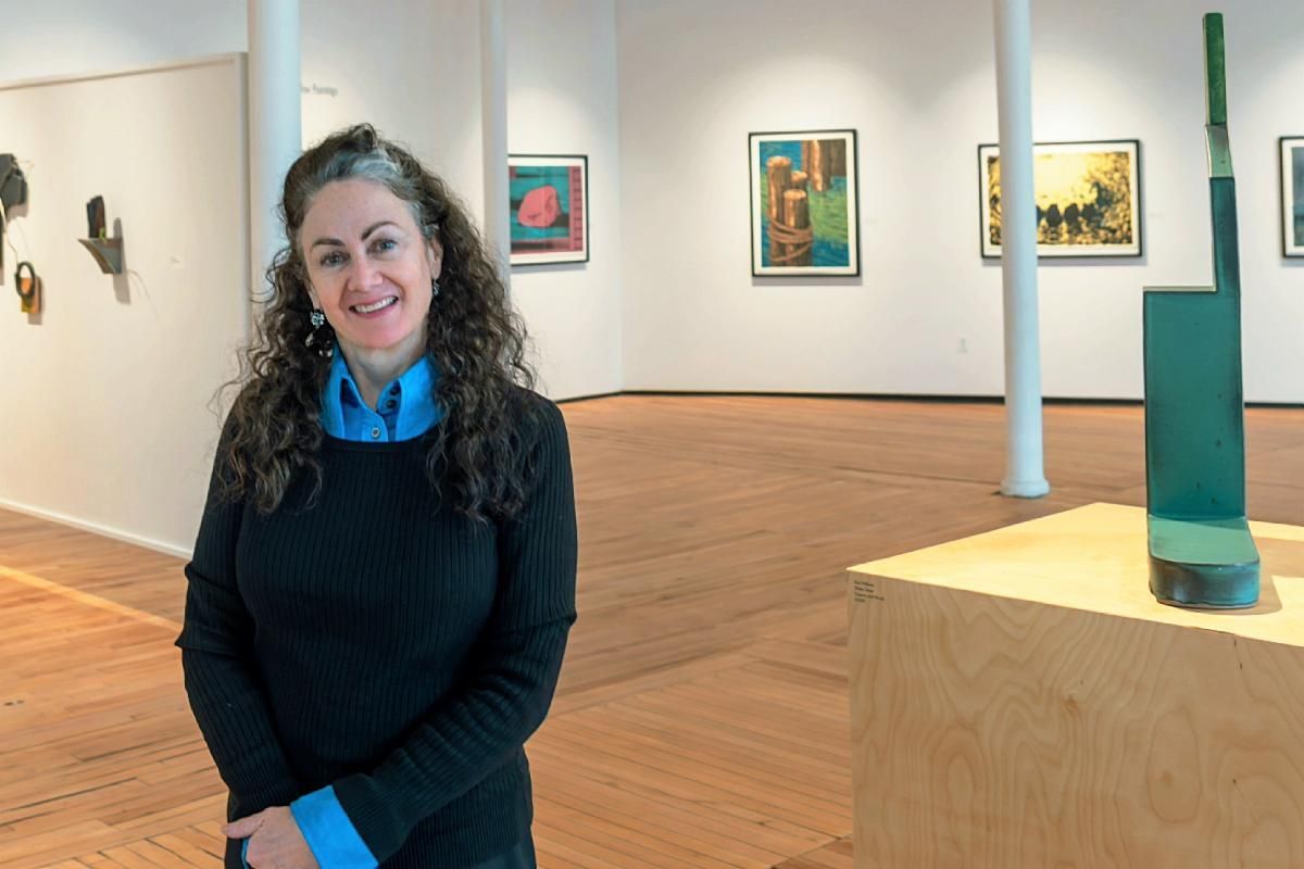 A smiling person in a blue shirt and black sweater stands in an art gallery featuring framed paintings and sculptures.