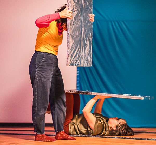 Two people in a studio; one stands holding a silver board, while the other lies on the floor supporting another board.