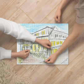Two people assemble a jigsaw puzzle of a sketch showing a building with yellow windows on a wooden floor.
