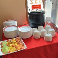 Buffet table with soup pot, fruit platter, plates, and cups on a red tablecloth.