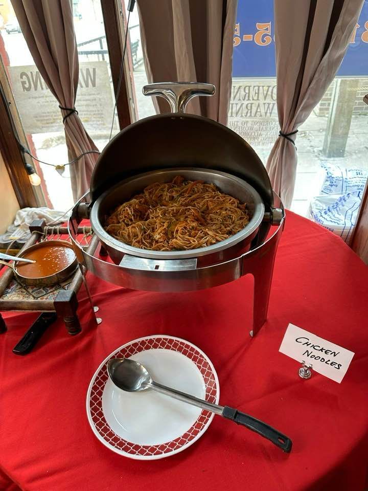 Chicken noodles in a chafing dish on a red tablecloth, with a side of sauce.