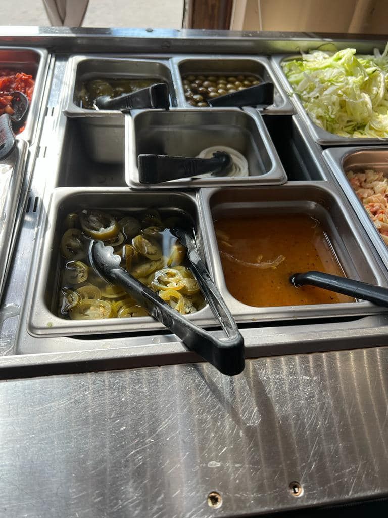 Salad bar with various toppings, including peppers, olives, lettuce, and dressing in stainless steel containers.