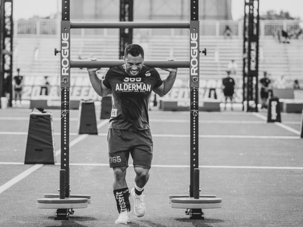 Athlete in a CrossFit competition carries a weighted bar, walking forward with a determined expression.