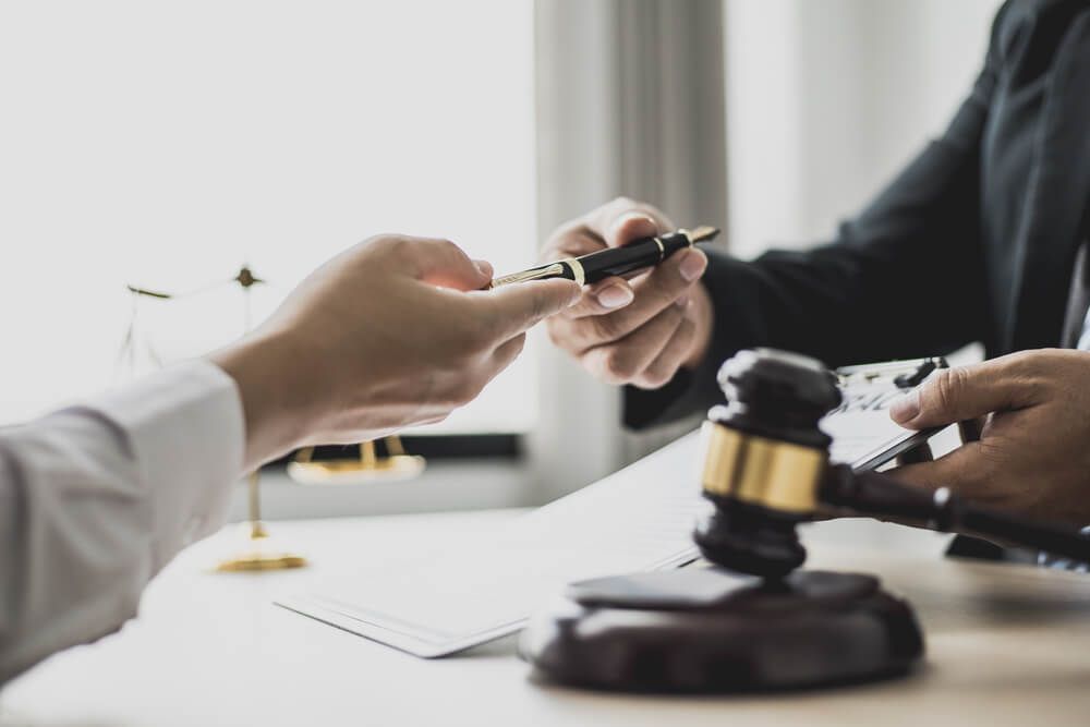 A Man is Handing a Pen to Another Man While Sitting at a Table — Morris Legal & Conveyancing in Forster, NSW
