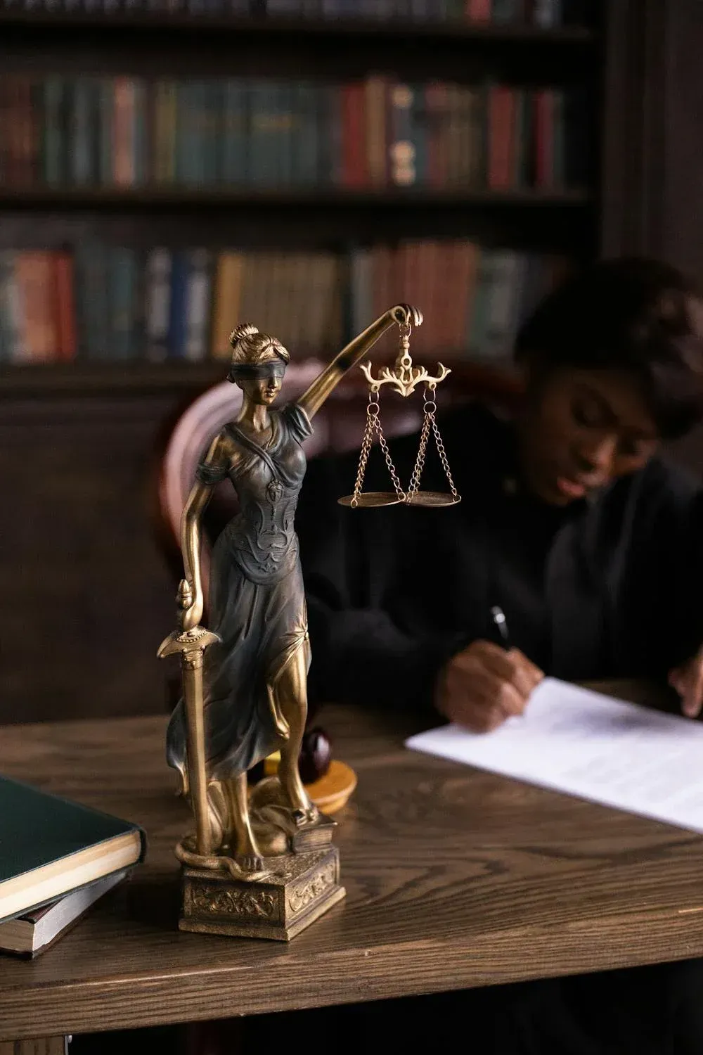 A statue of justice is sitting on a wooden table next to a man writing on a piece of paper.