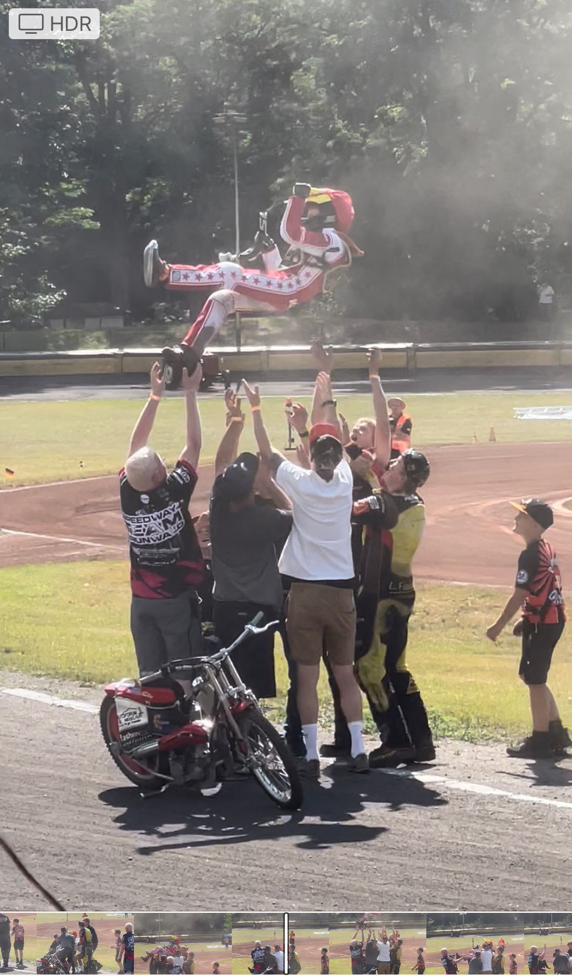 Ein Mann auf einem Motorrad wird von einer Gruppe Menschen in die Luft gehoben.