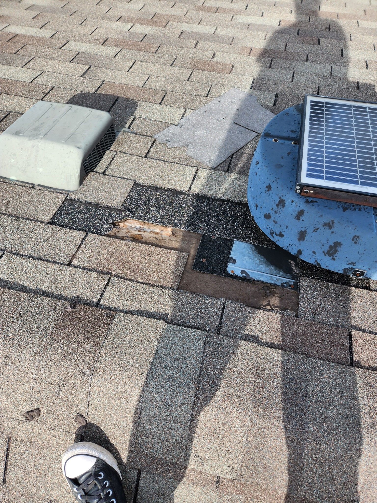 Rooftop with solar vent and wet shingles, showing a dark patch of roofing repair and a shoe at the bottom edge