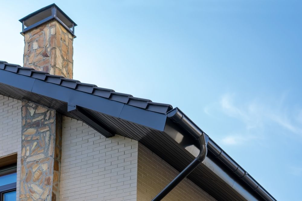 Black roof and gutter on a beige brick house, stone chimney against a blue sky.