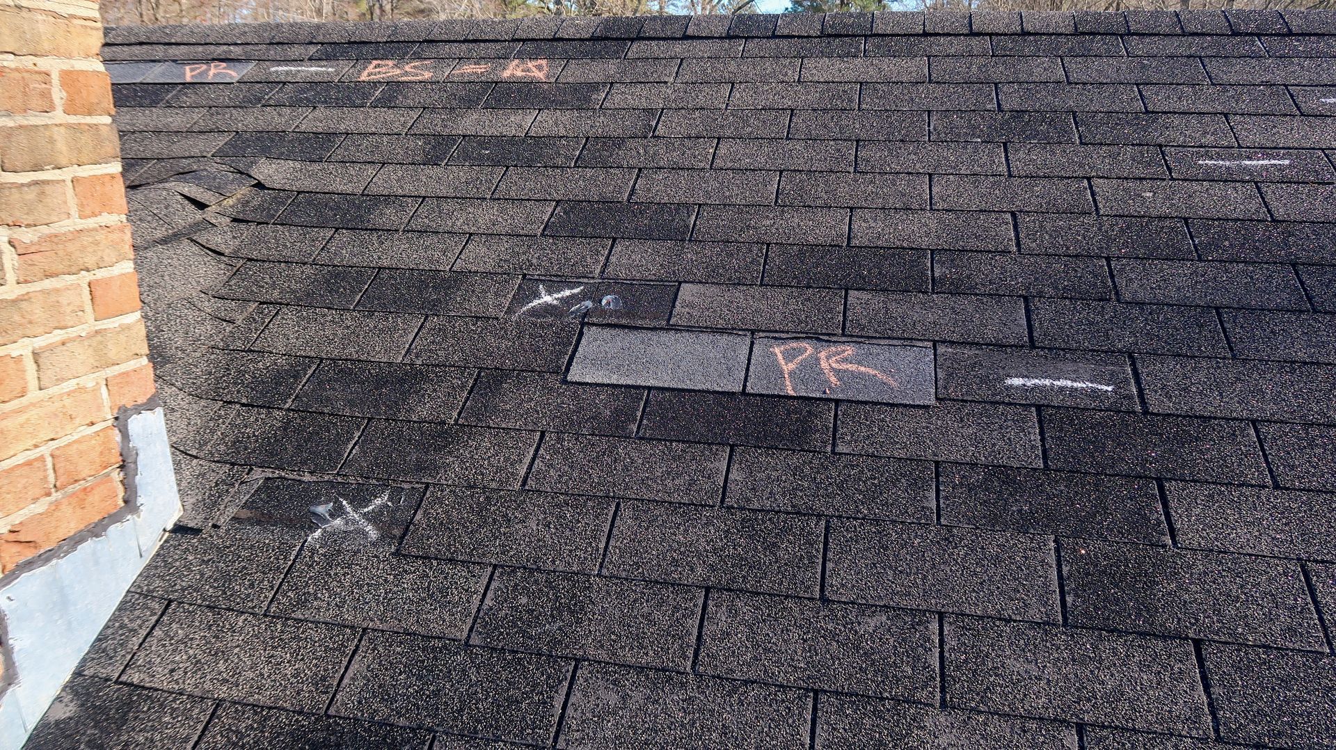 Damaged asphalt shingle roof with a patch near the center beside a brick wall