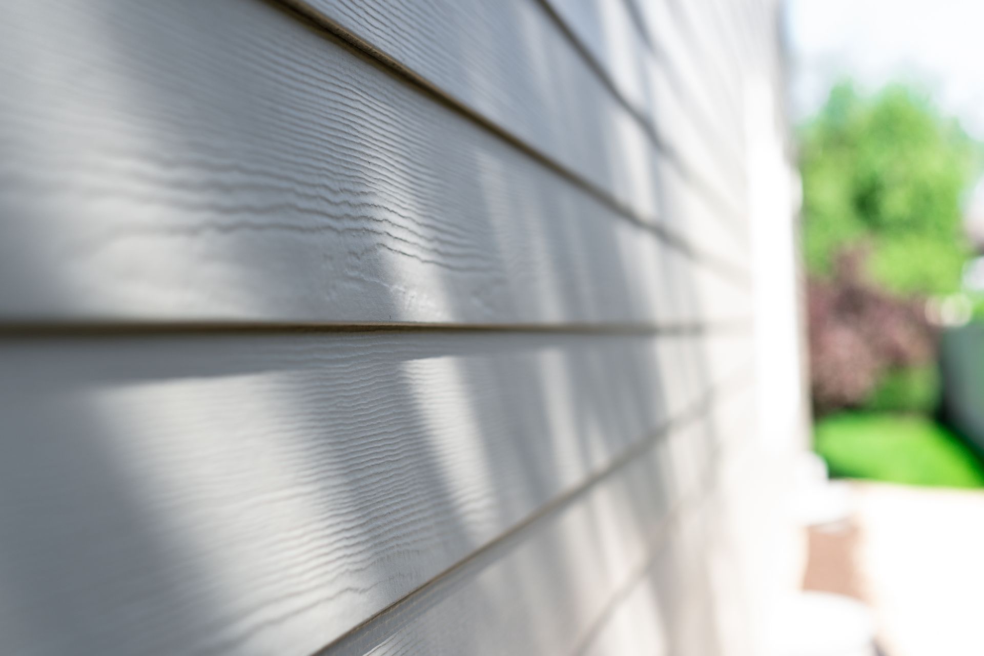 Close-up of white vinyl house siding with sunlight and a blurred green yard in the background