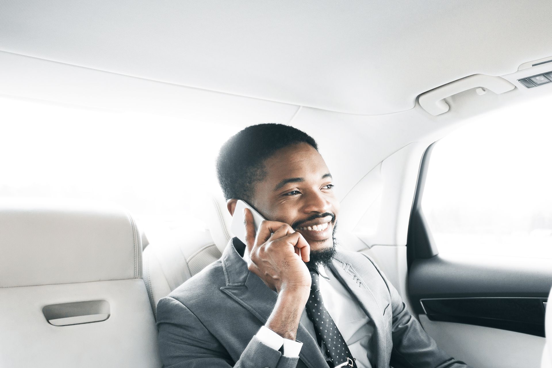 Man in a suit smiles while talking on a phone inside a car.