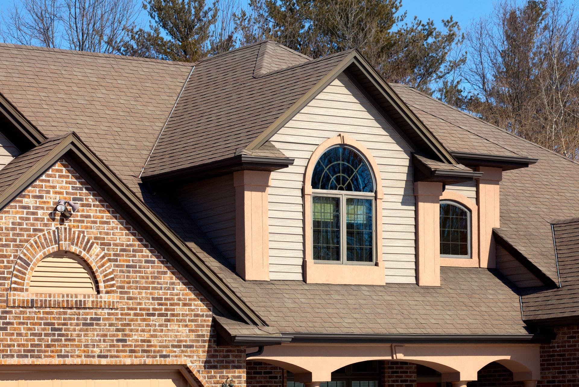 Brick house roof with tan shingles and arched windows against a blue sky
