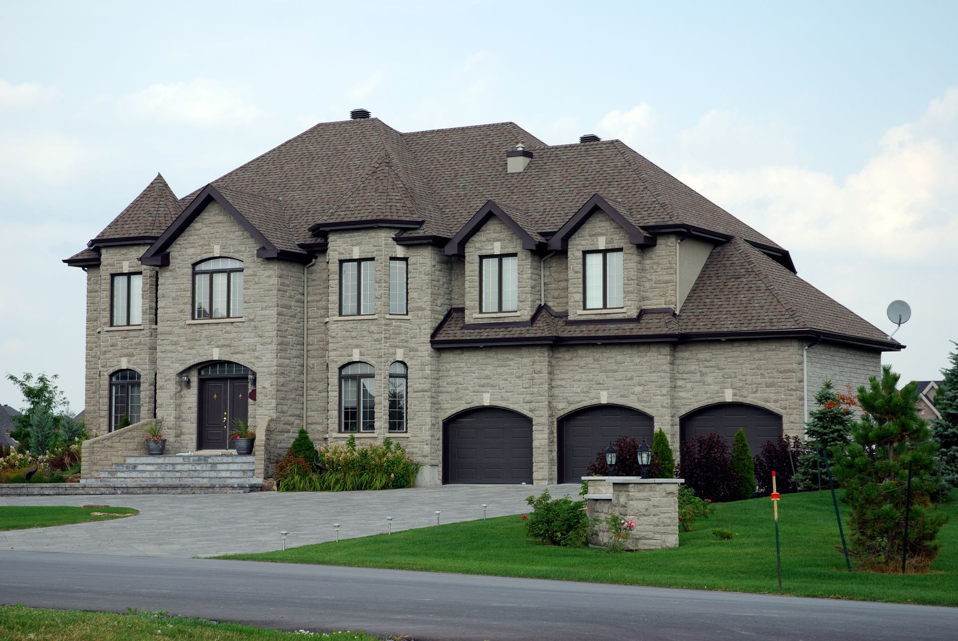 Large stone suburban house with a dark roof, three garage doors, and a landscaped front yard