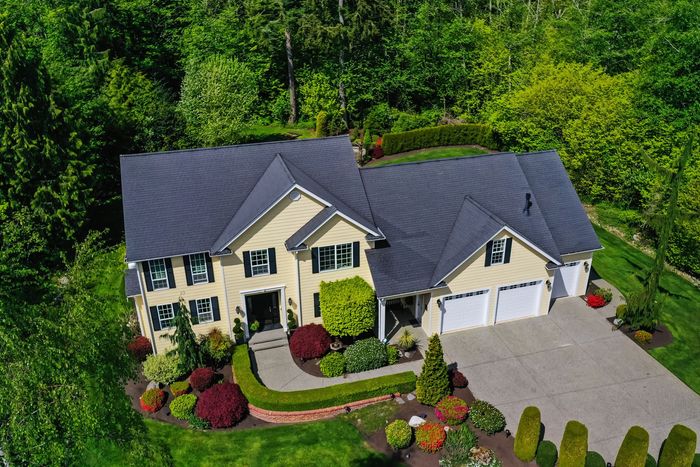 Aerial view of a beige house with dark roof, driveway, and landscaped yard surrounded by trees.