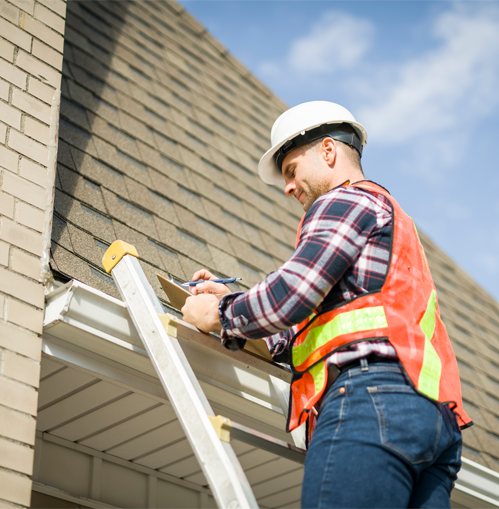Worker in hard hat and safety vest using a tool on a ladder beside a house roof