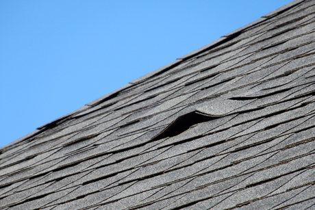 Gray shingled roof with a damaged section against a blue sky