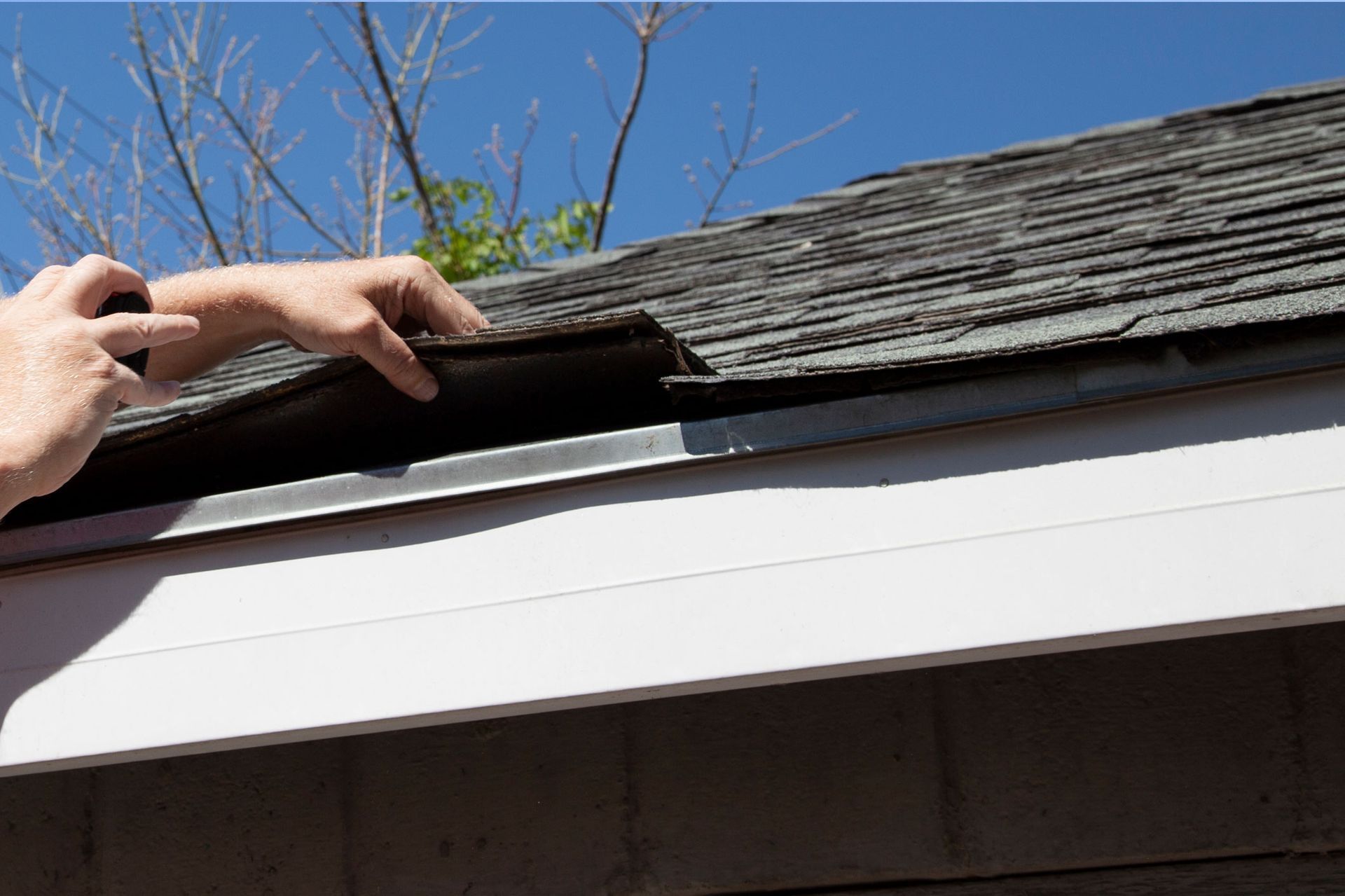 Hand inspecting roof shingles above white eaves under a blue sky