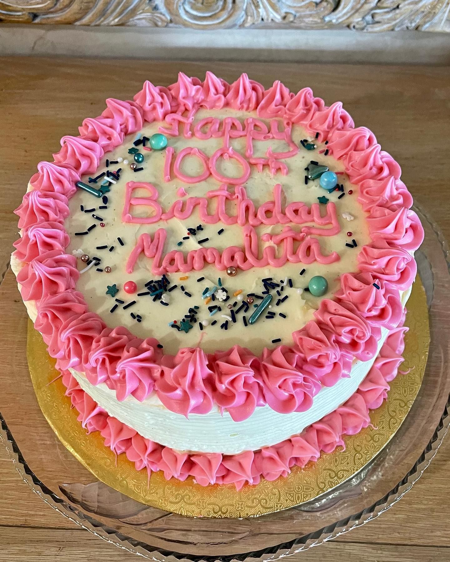 Pink-frosted birthday cake with “100 Birthday Nayel” written on top, on a gold cake board