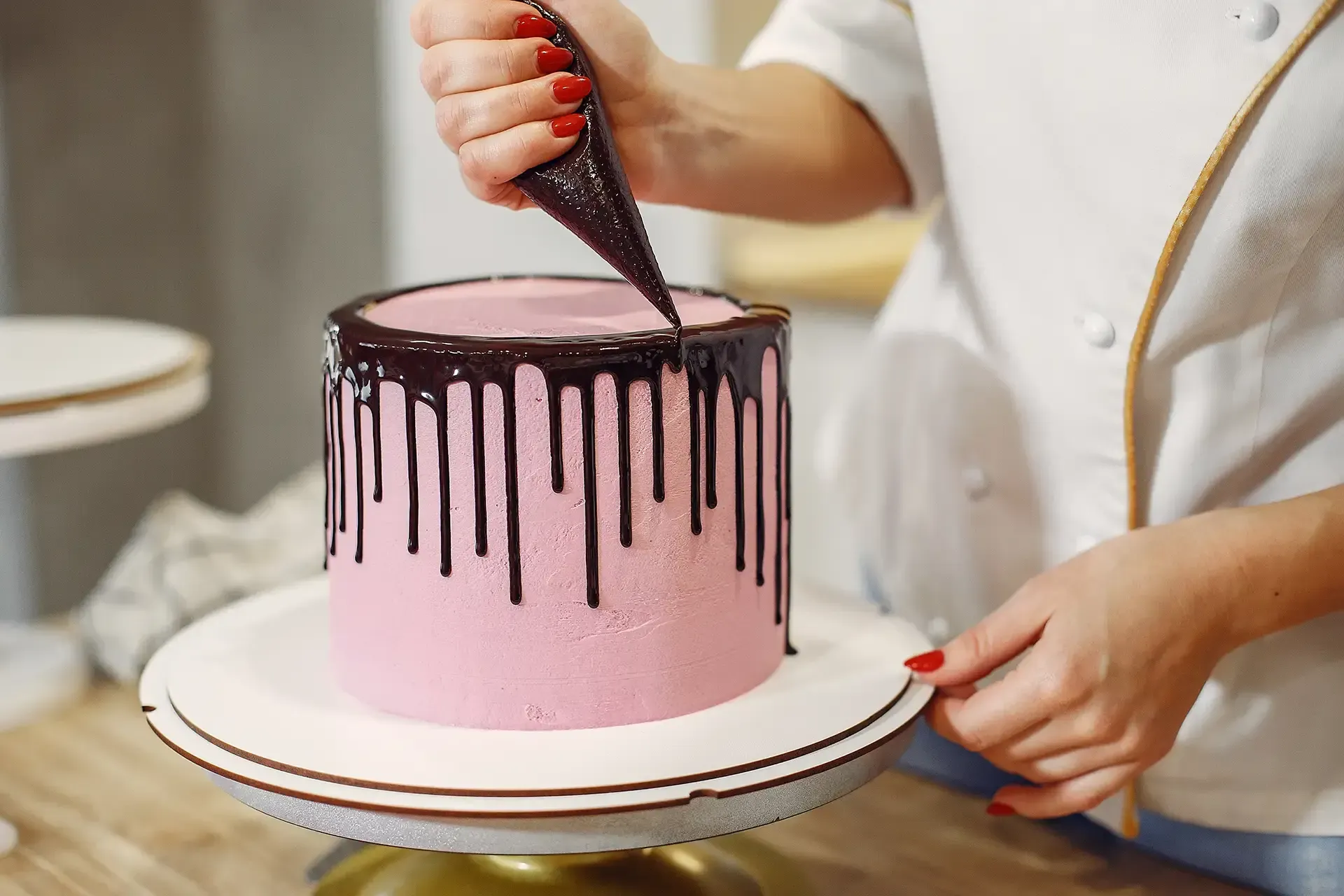 Decorating a pink drip cake with dark chocolate icing on a white stand