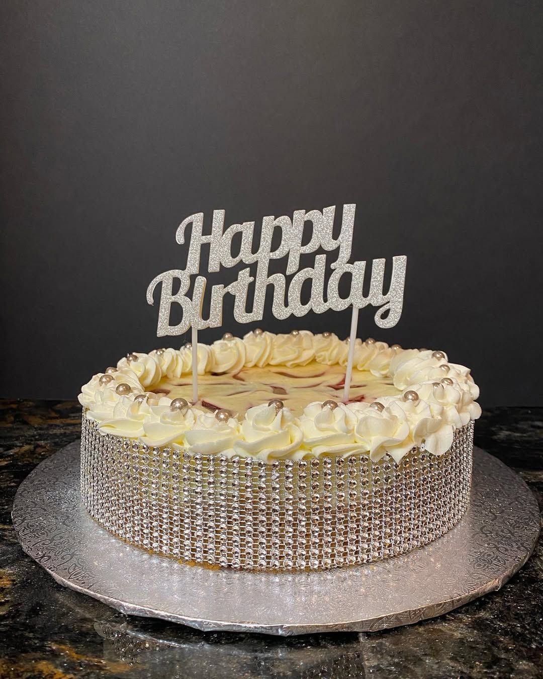 Birthday cake with gold sprinkles and a “Happy Birthday” topper on a silver cake board