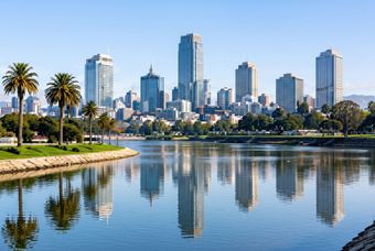 City skyline with tall buildings reflected in a calm lake, framed by palm trees and a grassy shoreline.