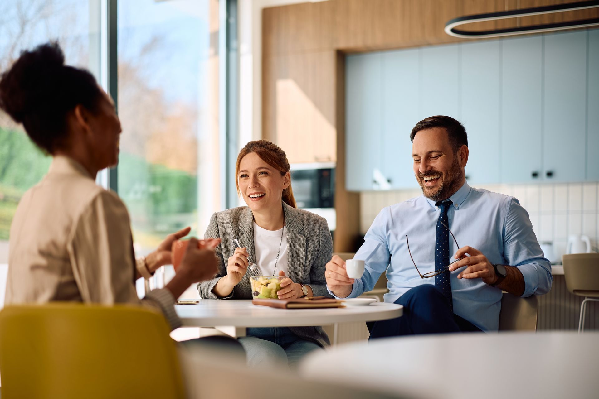 Three people chatting over drinks at a bright kitchen table, smiling in a modern home