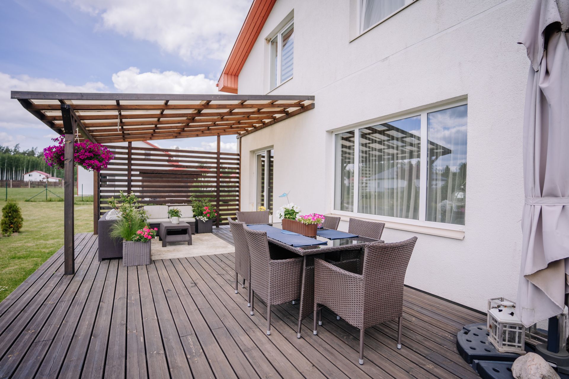 Modern patio deck beside a white house, with outdoor dining tables, wicker chairs, and a covered pergola.