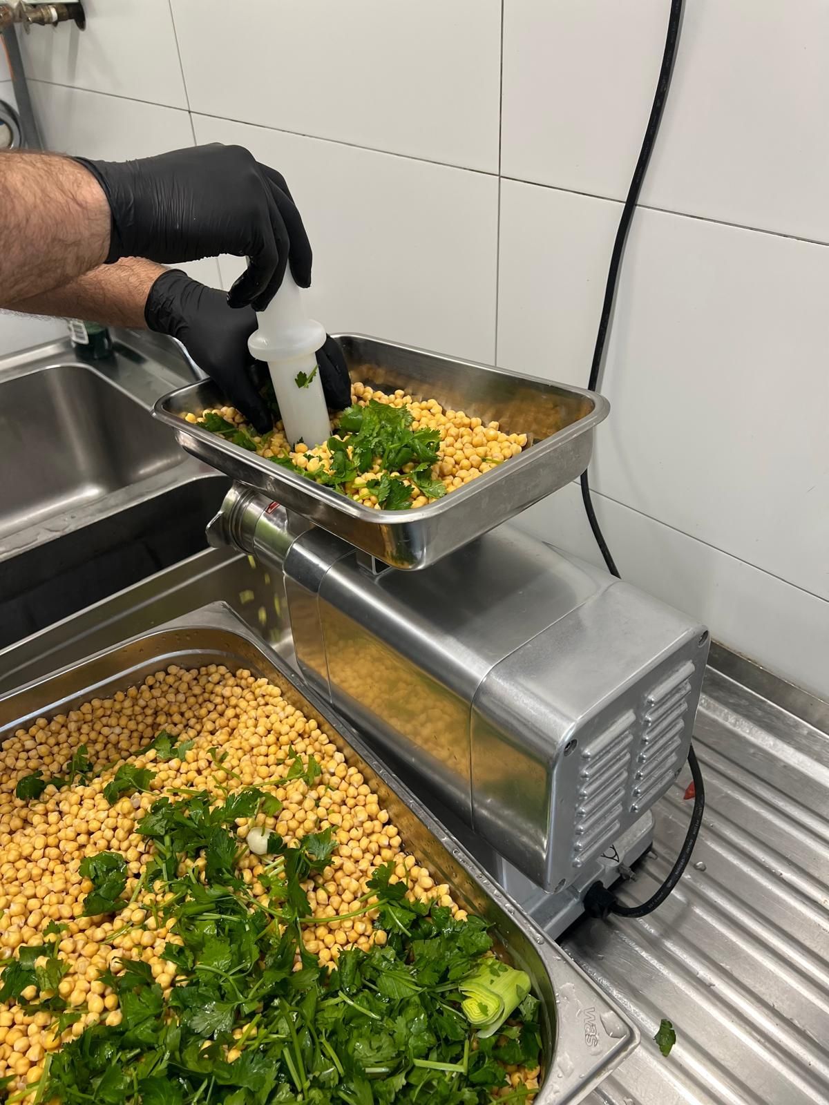 A chef in black gloves adds ingredients to a grinder, preparing food in a commercial kitchen.