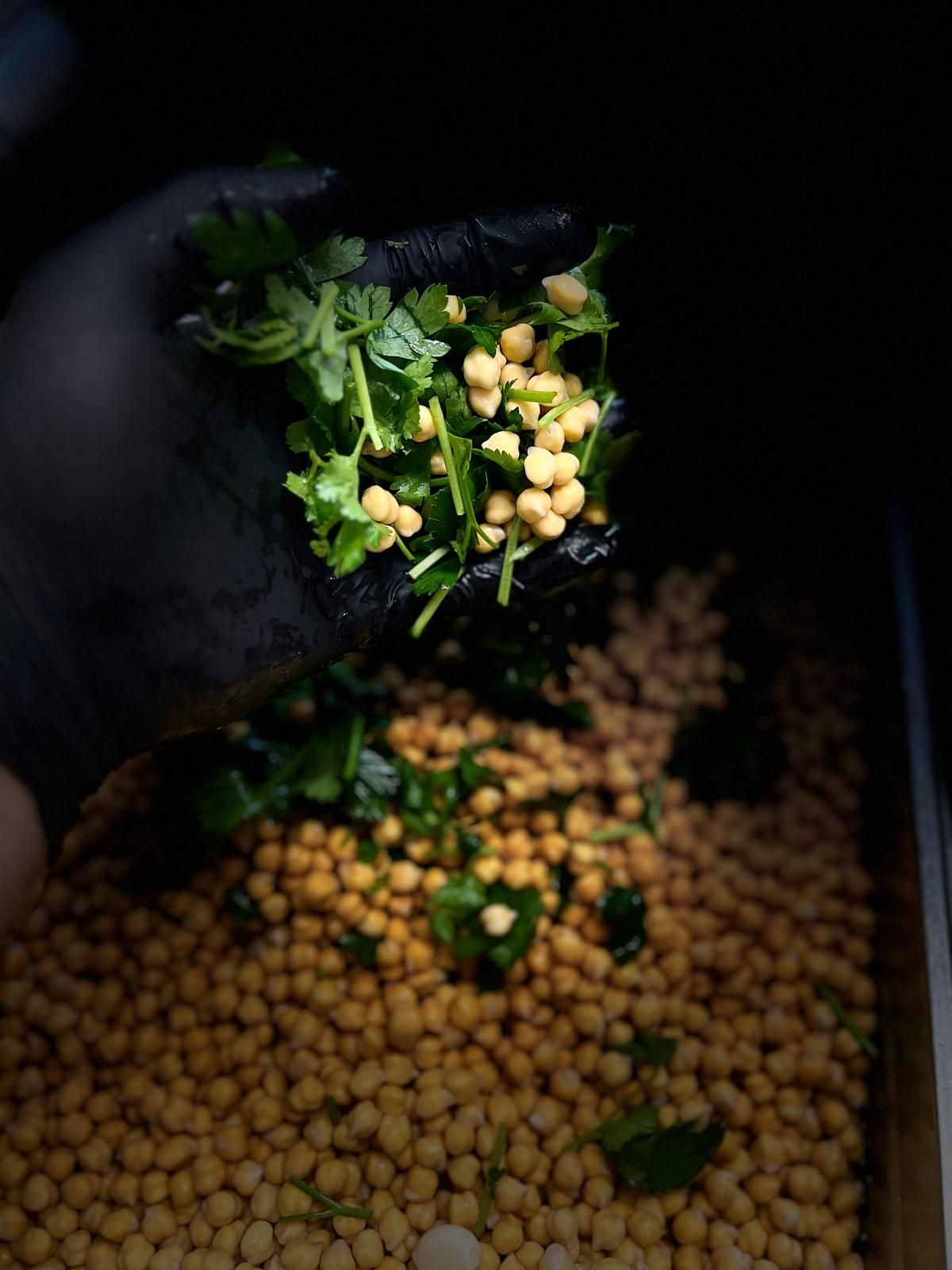Black-gloved hand adding chopped cilantro and chickpeas to a container of chickpeas.