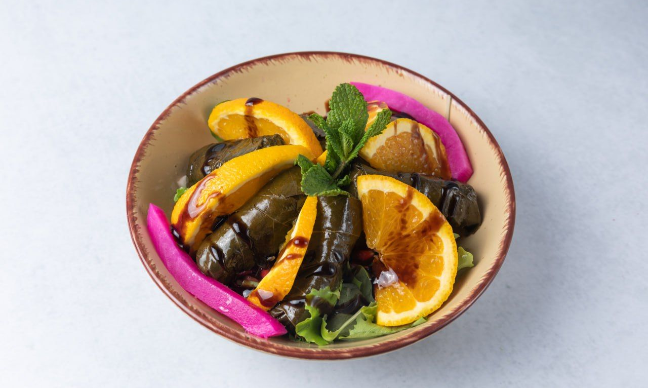 Bowl of stuffed grape leaves, orange slices, pink pickles, and mint sprigs; drizzled with sauce.