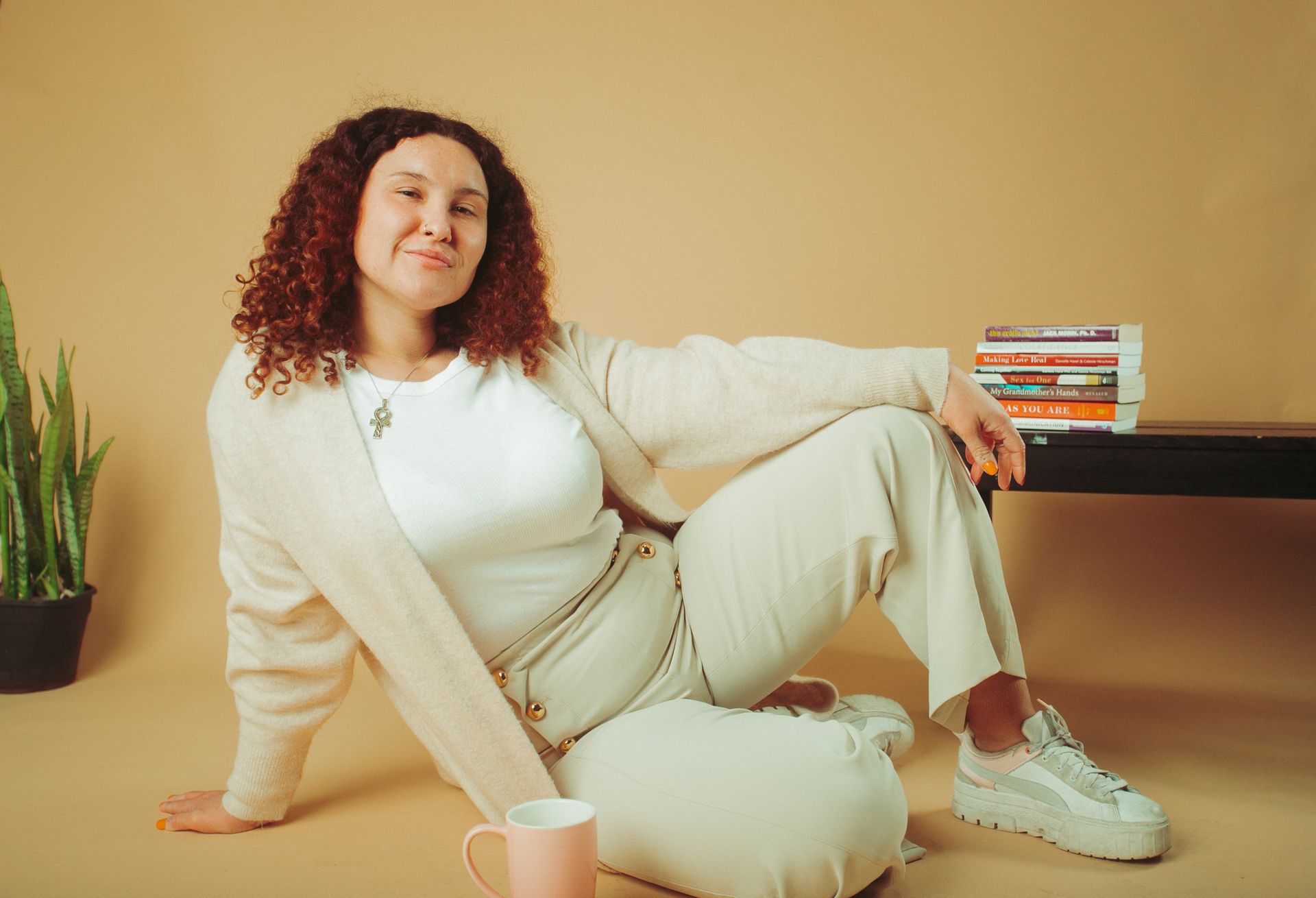 Woman in cream outfit, seated, smiles. Beside her is a mug, pillow, books and a plant against a tan background.