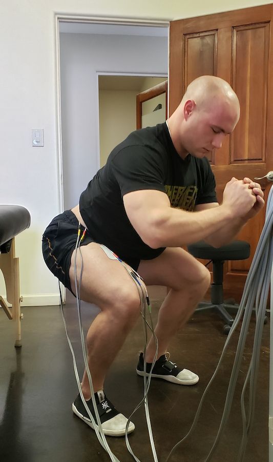 Man in a black shirt doing resistance-band squats in a home room.