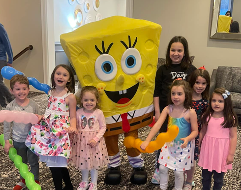 SpongeBob mascot posing with six children at a birthday party indoors.