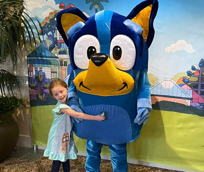 Child hugging a large blue cartoon dog character in front of a colorful mural