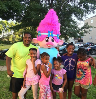 Five smiling children pose with a colorful pink troll mascot outdoors at a children'