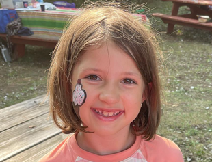 Smiling child with a face sticker, standing outdoors in sunlight near picnic tables and a wooden deck at a children's party. 
