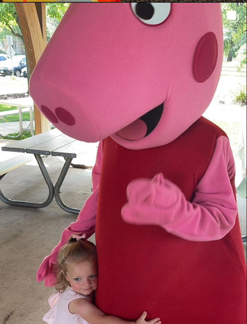 Pink dinosaur mascot hugging a small child outdoors near a picnic table