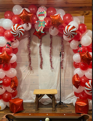 Red, white, and silver balloon arch with holiday elf topper and small stool beneath it at a party. 