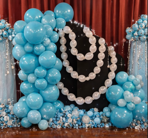 Blue and white balloon arch with white pearl balloon garlands on a black backdrop and red curtains at a party. 