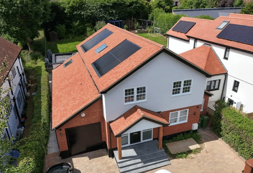 Two-story house with red roof tiles and black solar panels, white and brick exterior, surrounded by green foliage.